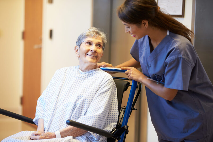 Female porter pushed an elderly female patient in a wheel chair whilst looking at each other and smiling.