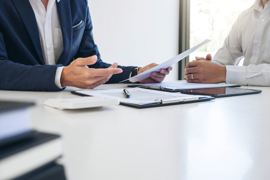 Two men sit at a table facing each other and reviewing paperwork.