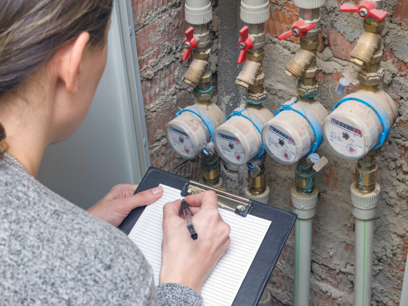 A woman crouches down in front of water meters to take a reading and is holding a clipboard and pen.